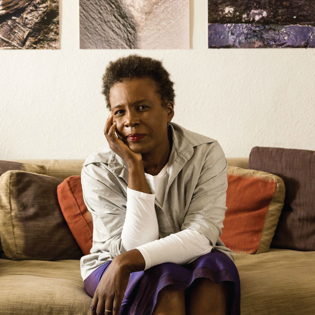 Photo of author Claudia Rankine sitting on a couch in front of artwork.