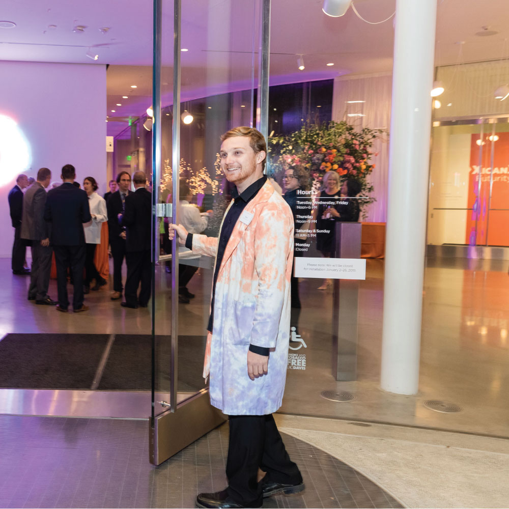 Student in tie-dyed lab coat opening museum door for visitors.