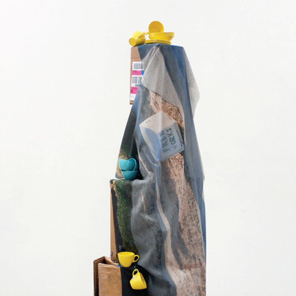 Sculpture with cloth draped over boxes and colorful plasticware stacked on it.