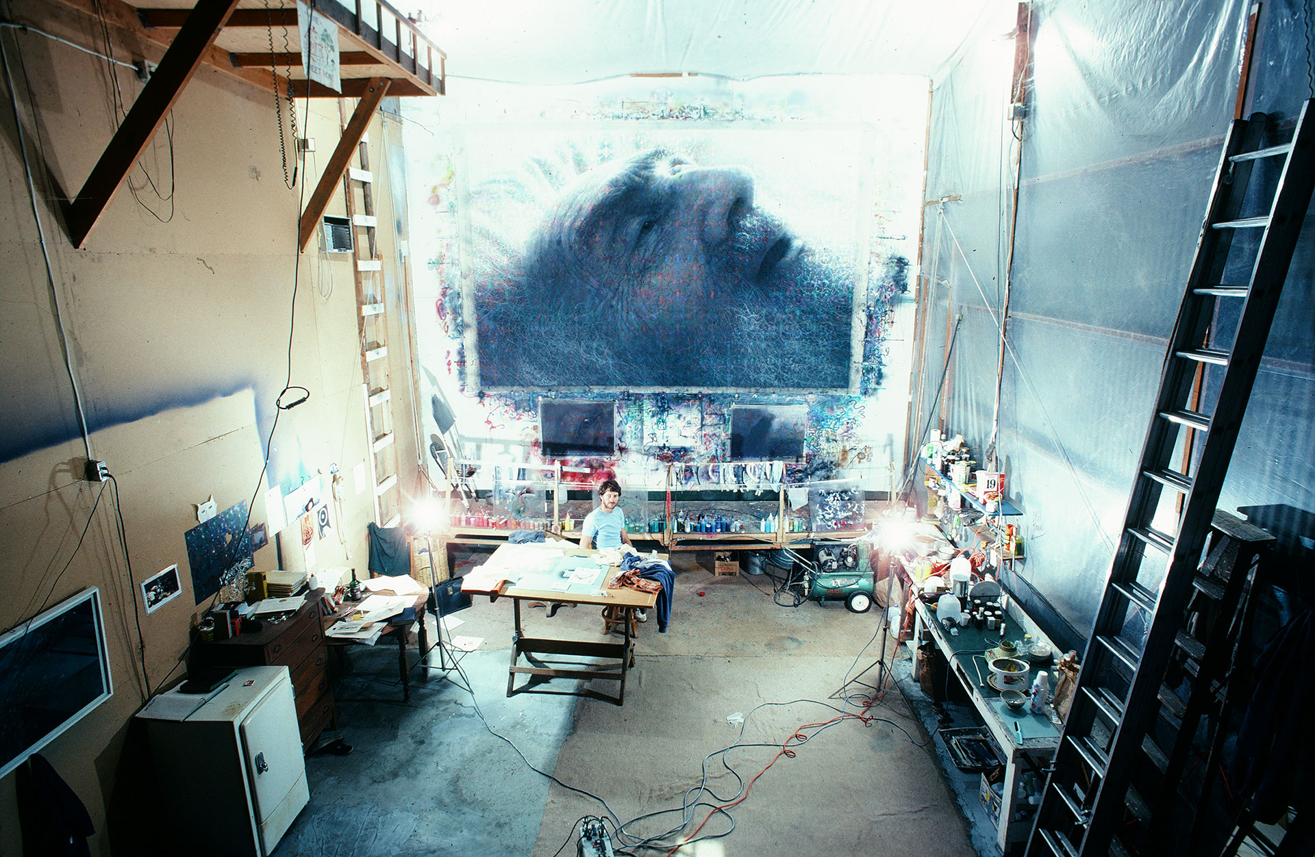 Photo of man in art studio with large painting of elderly man in the background.