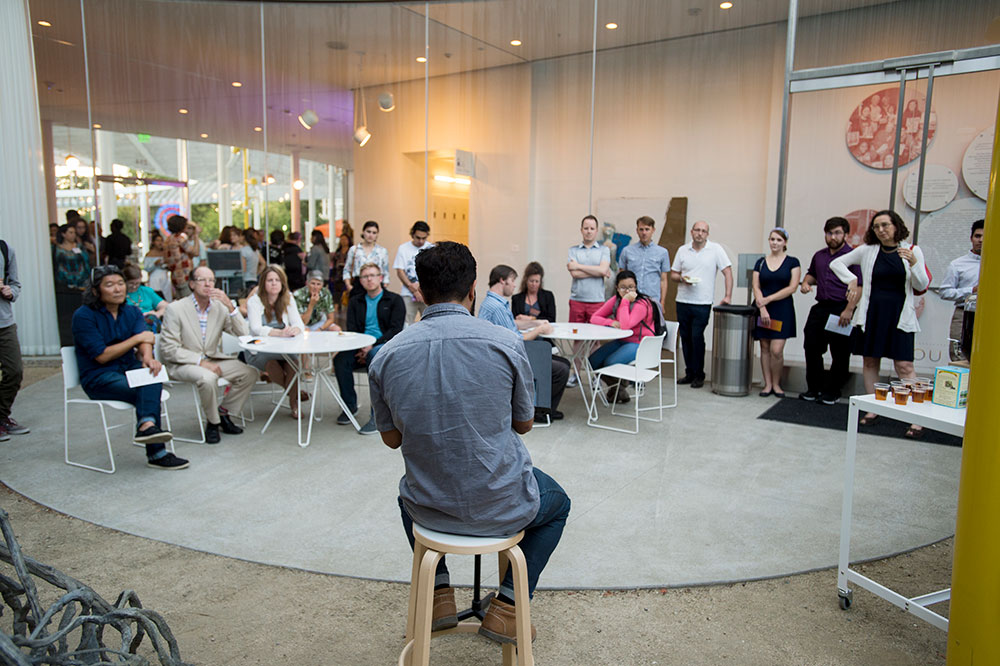 Crowd of people seated in courtyard listening to a speaker seated on a stool in the foreground.