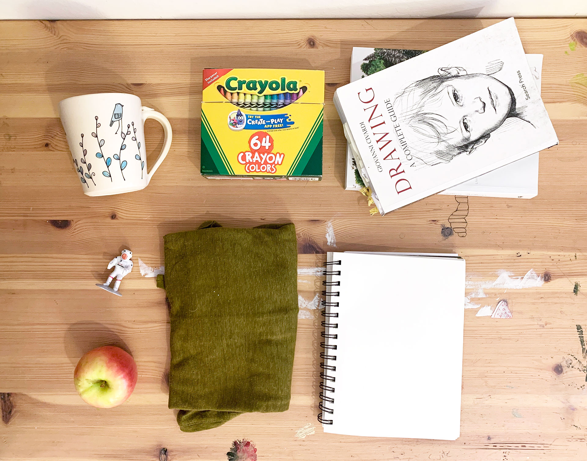 Art materials, mug, plastic toy, books, and apple on a wood table.