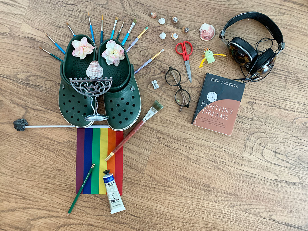 Various found objects like shoes, headphones, flag, shells and art supplies on a wooden table.