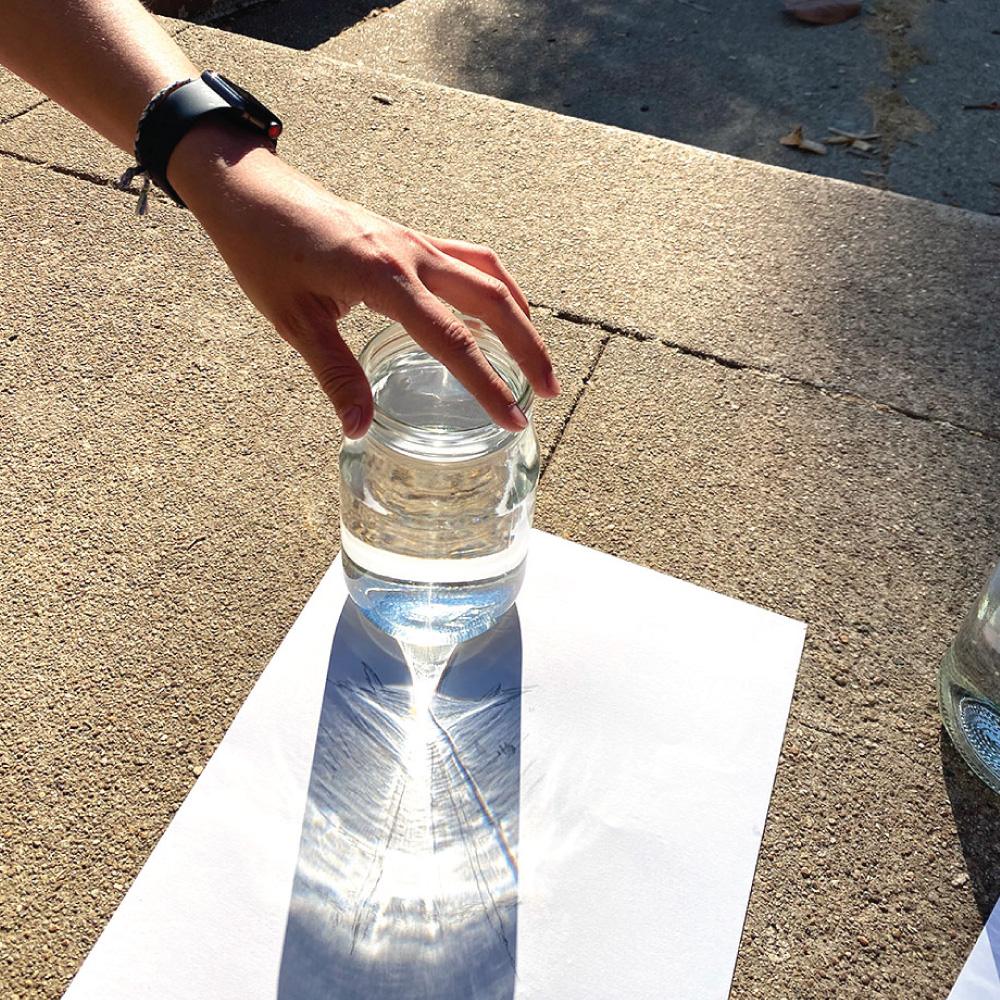 Hand placing water filled glass on paper to cast shadows and light from the sun.