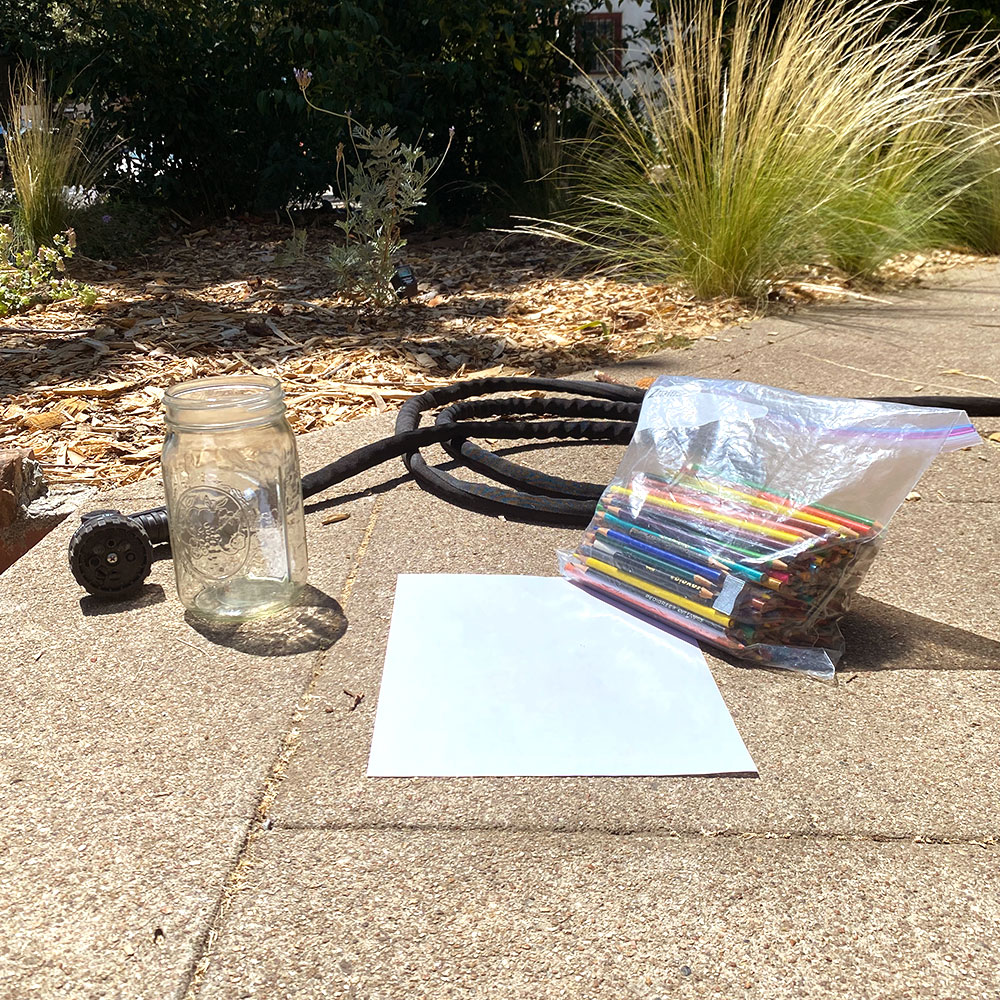 Empty glass, white paper, bag of colored pencils and garden hose arranged on sidewalk in the sun.