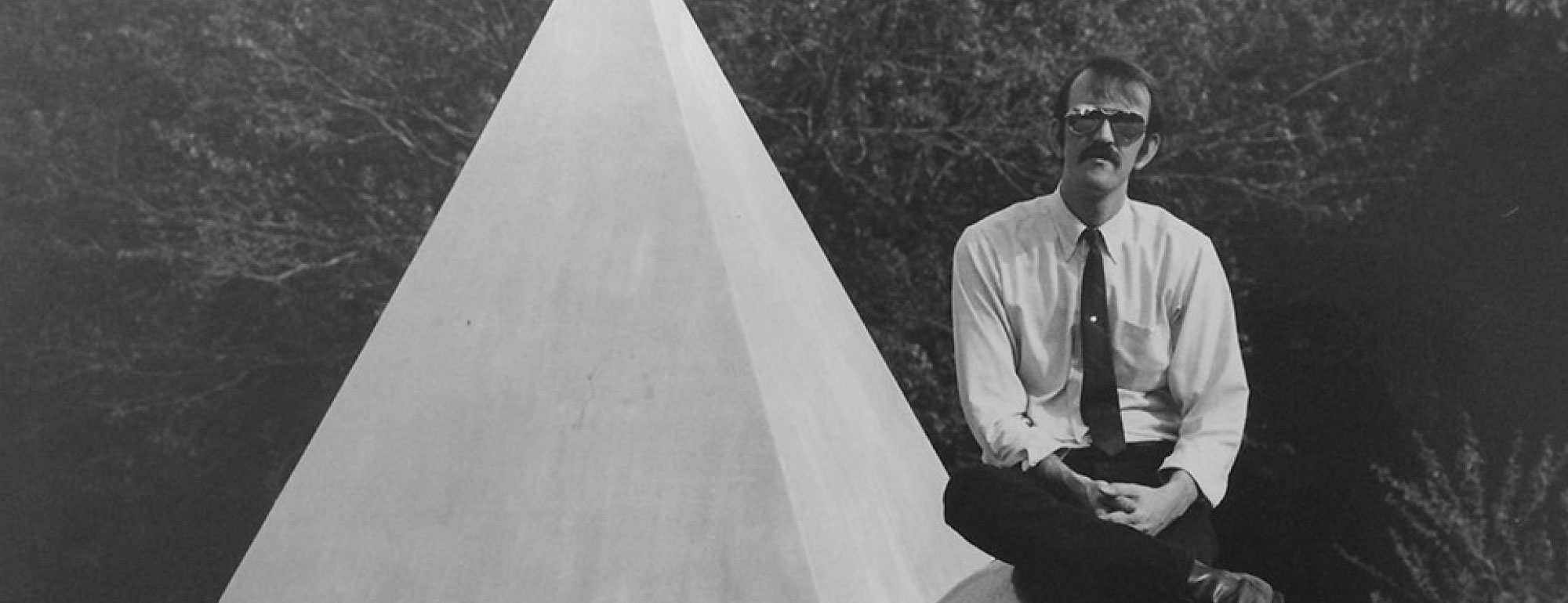 Black and white photo of man sitting on ball chained to pyramid.