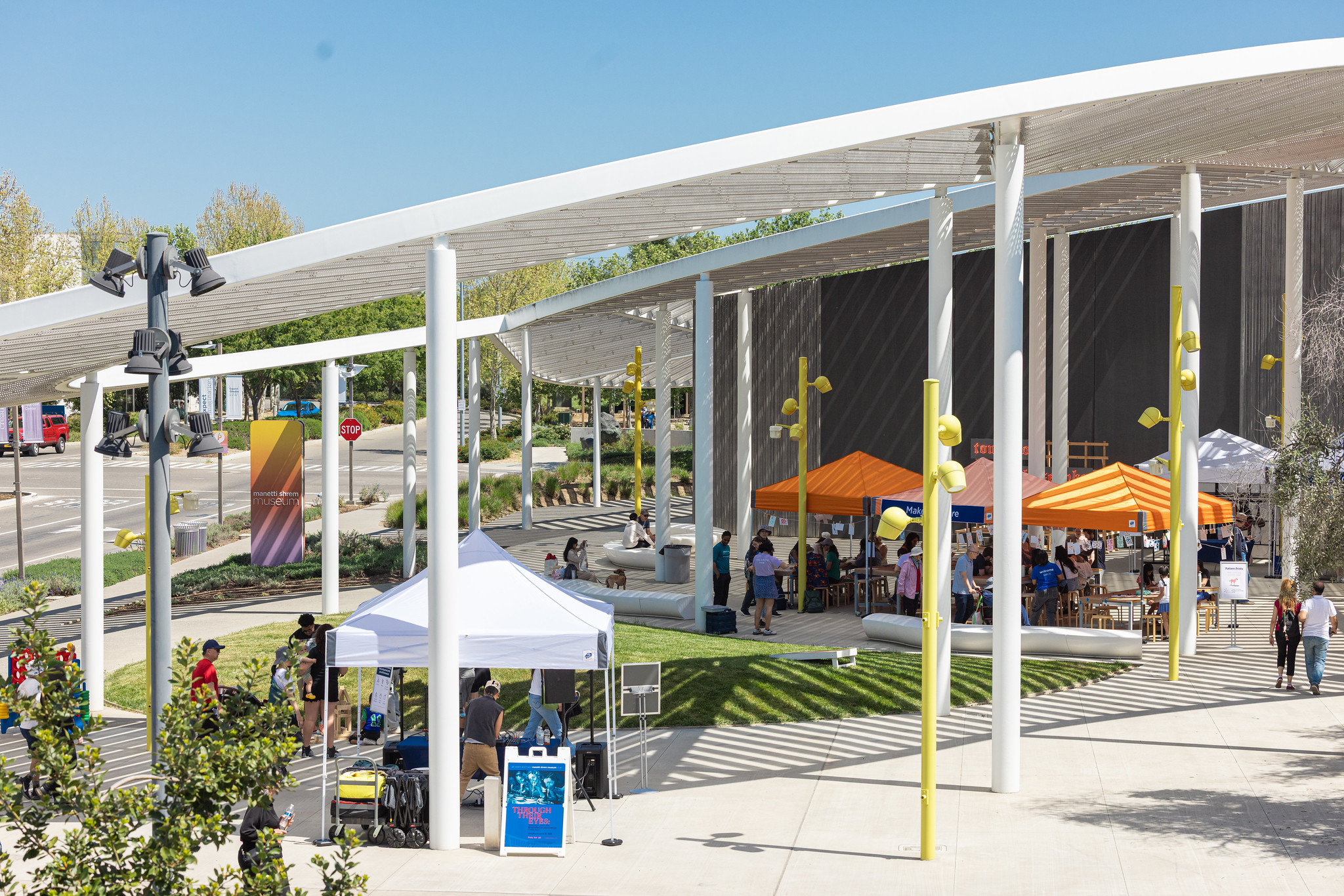 Groups of people gather under orange and white tents on the plaza of a museum under a swooping white canopy.