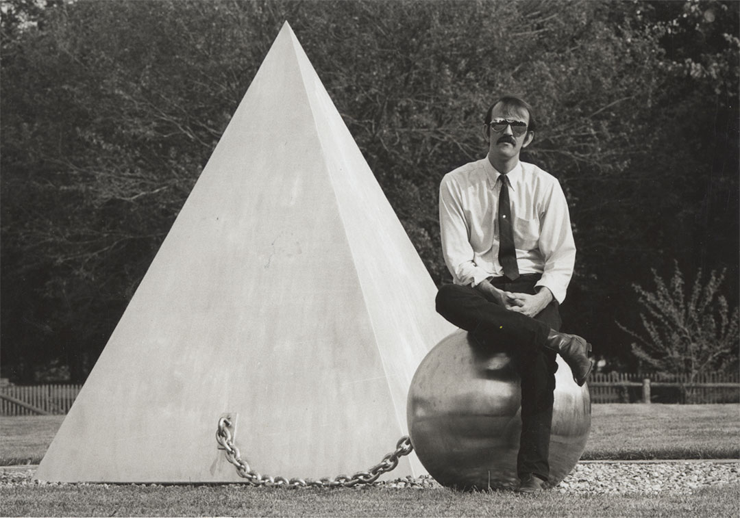 William T. Wiley sitting on a ball in front of a pyramid.