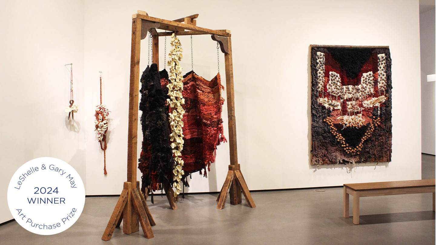 wo woven tapestries made with chains, twisted rope, ceramics and woodA selection of works by April Camlin, MFA, winner of the LeShelle & Gary May Art Purchase Prize, from left to right: It’s Coming Back, Spirit House and Where They Broke Me, 2024. and in shades of burgundy, black and ivory are displayed in a gallery. One hangs from a wooden structure, and the other is mounted on the wall.