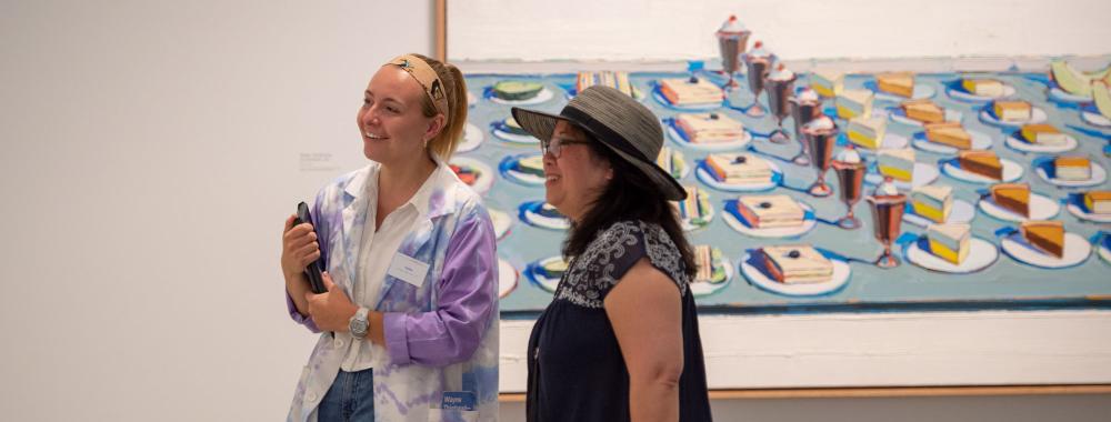 Student staff and visitor looking at art in the gallery with a Thiebaud painting of pies behind them.