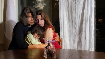 Two people and a child hugging while sitting at a table with a window behind them.