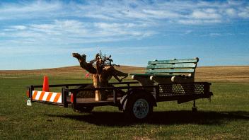 Transplant Module, 2021, 35mm film series. Utility trailer; replica of an NYC park bench; lodgepole pine roots; soil from a construction site; traffic cone; road barrier; survey flags; open prairie.