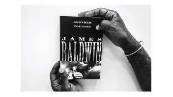 Black and white photo of hands holding a book titled 'Another Country' by James Baldwin.