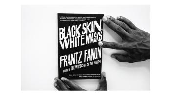 Black and white photo of hands holding a book titled 'Black Skin, White Masks' by Franz Fanon.