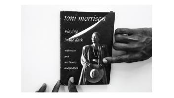 Black and white photo of hands holding a book titled 'Playing in the Dark' by Toni Morrison