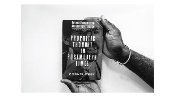 Black and white photo of hands holding a book titled 'Prophetic Thought in Postmodern Times' by Cornel West.