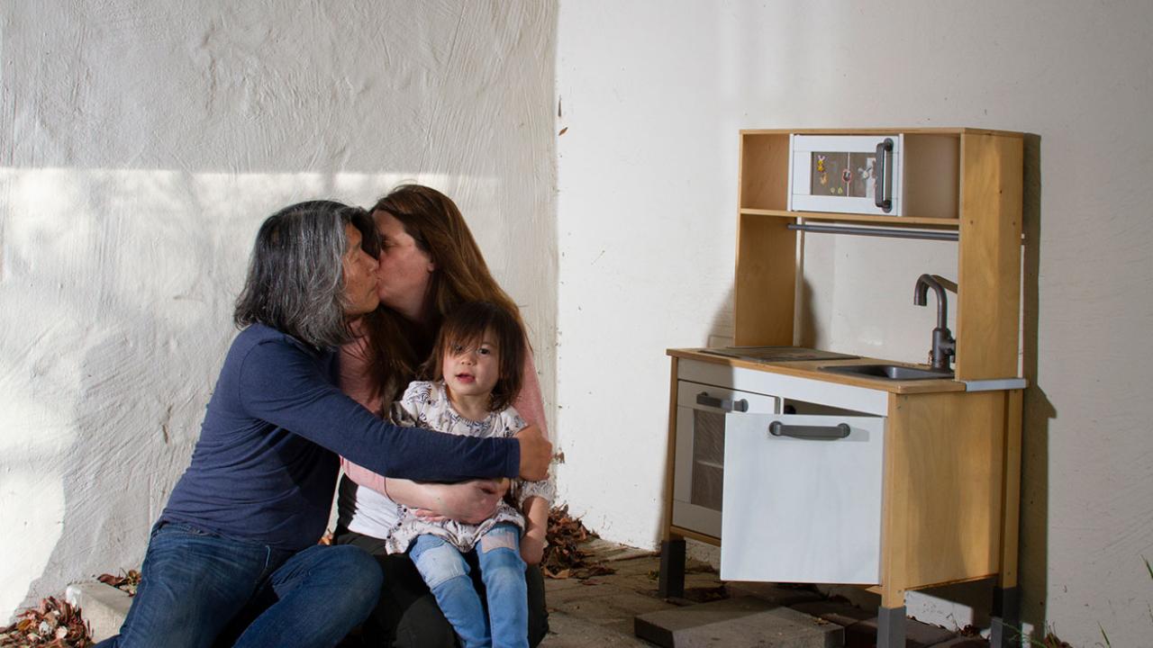 Two people and a child hugging in front of a white wall.