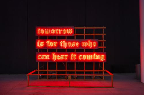 Red neon sign on a wood frame in front of the museum. Text reads 'Tomorrow is for those who can hear it coming'.
