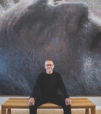 Artist Stephen Kaltenbach sitting in front of his painting 'Portrait of My Father' showing an elderly man's face lying down.