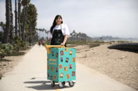 Artist Ariana Arriaga standing outside with a small green painted cart on wheels covered in stickers she created.