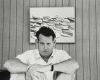Black and white photo of Wayne Thiebaud sitting in front of one of his Pies paintings.