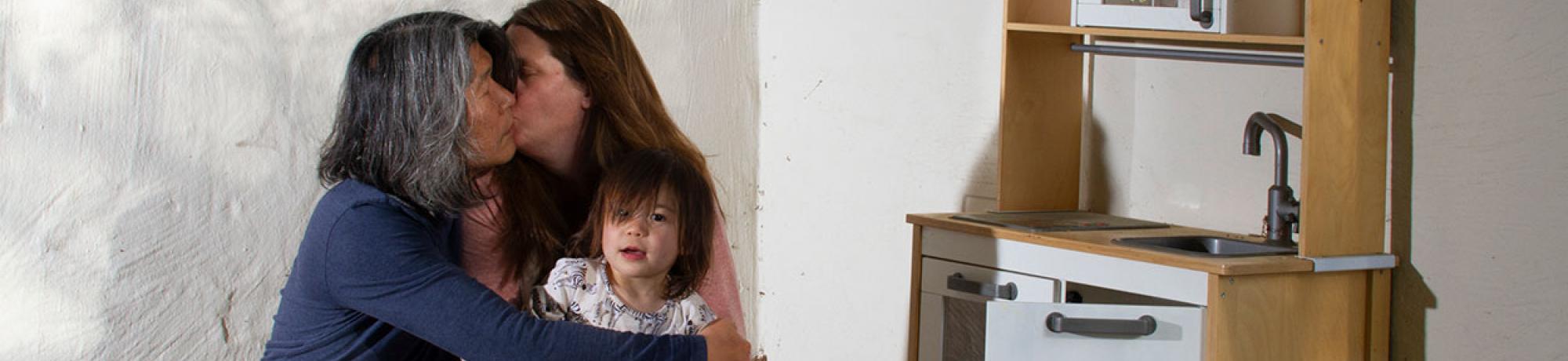 Two people and a child hugging in front of a white wall.