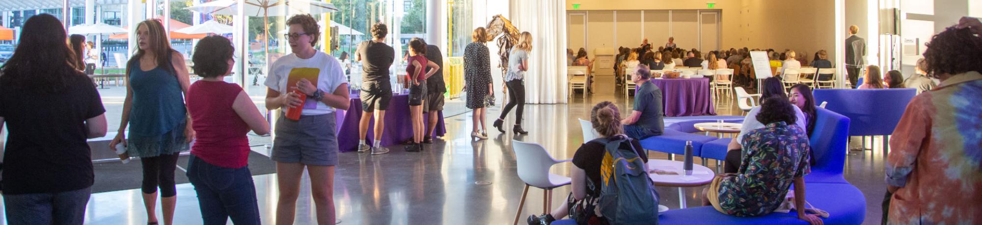 Visitors talking in the museum lobby while a program goes on in the background with an audience and artists talking.