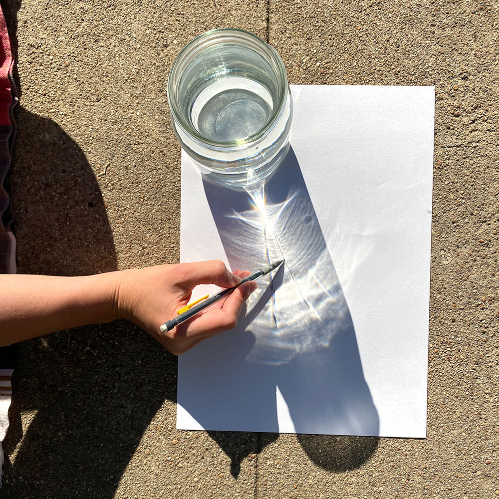Using a pencil to sketch the shapes created by the light and shadows of the sun shining through the water filled glass.