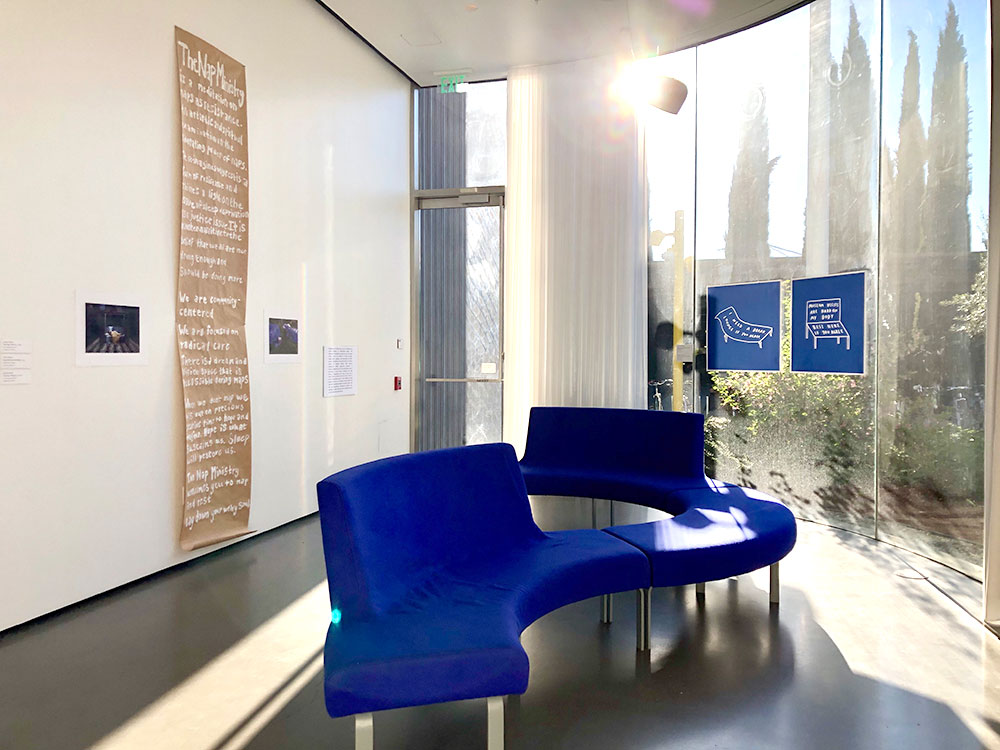 Brown paper banner with white painted text hanging on wall behind a blue winding couch. Window with two pieces of artwork on it.