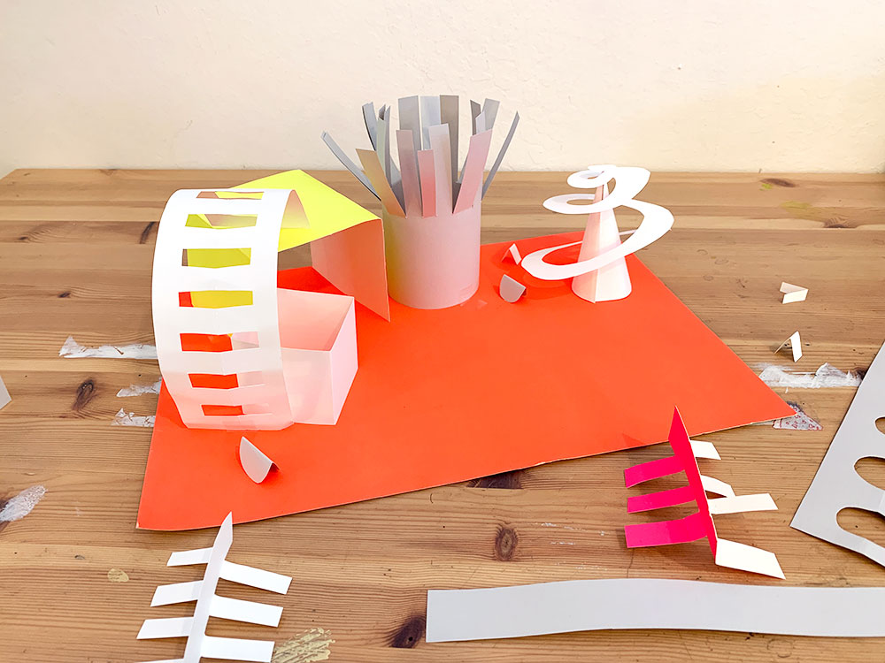 Brightly colored paper cut and folded into abstract shapes like cubes, tubes, and swirls.