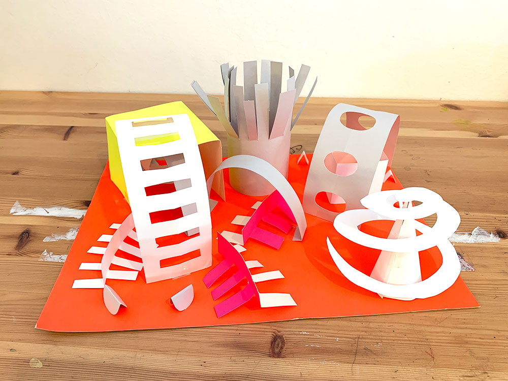 Brightly colored paper cut and folded into abstract shapes like cubes, tubes, and swirls.